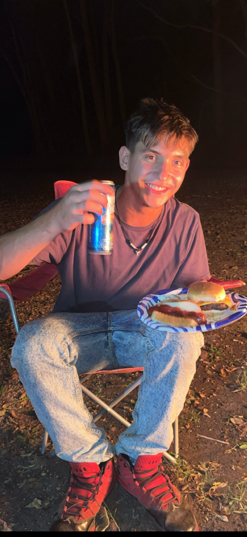 Man sitting outdoors at night, holding a can and a plate with food.